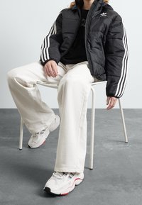 Chaqueta de plumas negra con rayas blancas, combinada con una camiseta negra, pantalones anchos blancos y zapatos deportivos gris-blanco con acentos rojos.