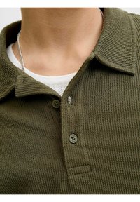 Polo shirt vert olive foncé en tissu texturé, avec un col, deux boutons et une poche sur le côté gauche.