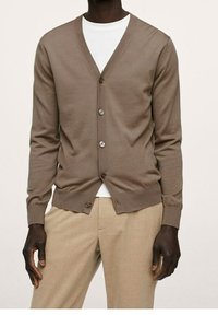 Cárdigan de punto marrón con escote en V, cinco botones marrones y puños acanalados. Combinado con una camisa blanca y pantalones beige.