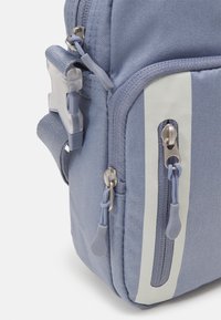 Modrý látkový vak s hladkou texturou, kapsami na zip, sportovními pruhy a plastovým kováním se šedými detaily.