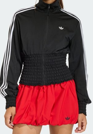 Kobieta w czarnej kurtce Adidas na zamek z białymi paskami i marszczonym talią, połączona z czerwoną spódnicą w stylu balonowym z logo Adidas.