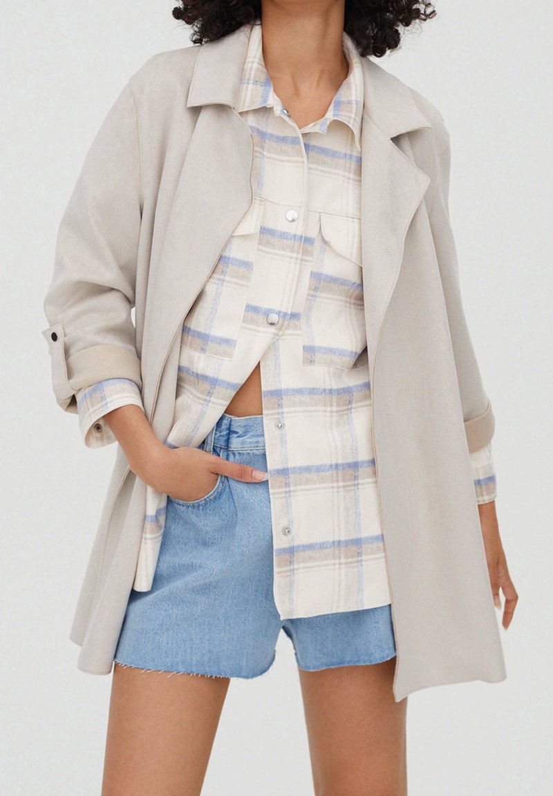 Beige ruskinds trenchcoat lagd over en lys plaidskjorte med blå og beige nuancer, parret med slidte denimshorts.