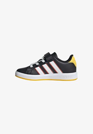 Chaussures de sport noires avec des rayures blanches et rouges, lacets argentés, bride en velcro, accent jaune au talon et semelle blanche. Matière synthétique lisse.