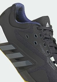 Chaussure de sport noire avec une tige en mesh et synthétique, motifs texturés, trois bandes réfléchissantes et un col rembourré. Semelle extérieure jaune.