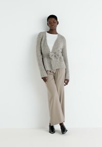 Cardigan texturat de culoare gri deschis, cu un nod în față, purtat peste un top alb. Asortat cu pantaloni largi de culoare crem și tocuri negre. Design simplu.