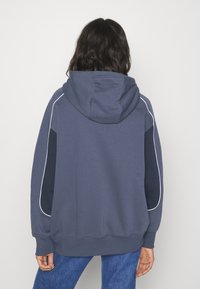 Hooded sweatshirt in dark blue with white piping details, featuring a relaxed fit and contrasting panels on the sides. Soft fabric texture.