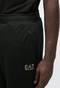 Melnas sporta bikses ar faktūrētu apdari, ar zelta "EA7 Emporio Armani" logotipu un sānu kabatu ar rāvējslēdzēju.