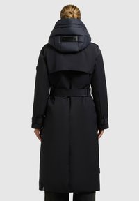 Lang zwarte pufferjas met een afneembare, oversized capuchon. Heeft een ceintuur in de taille en manchetten met banden. Soepele textuur met een matte afwerking.