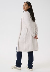 Cardigan beige chiaro realizzato in tessuto a maglia texturizzato, con un design lungo e aperto e sottili accenti luccicanti. Indossato sopra jeans scuri.