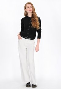 Zwarte gebreide cardigan met een textuurpatroon, ronde halslijn en knoopsluiting, gecombineerd met witte wijde broeken en zwarte schoenen.