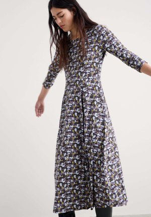 Hosszú sötét hajú nő, aki hosszú ujjú, bokáig érő virágmintás ruhát visel lila, fehér és sárga virágokkal, kinyújtott karokkal áll.