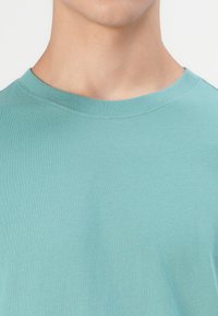 Tricou cu mânecă scurtă din țesătură teal deschis, cu guler rotund și o textură netedă, moale, fără modele sau accente vizibile.