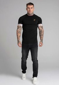 Juoda aptempta marškinėliai su baltais ir auksiniais akcentais; derinama su nudėvėtais tamsiais džinsais ir baltais sportbačiais. Matomos tatuiruotos rankos.