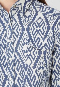 Camisa de botones hecha de un tejido texturizado con un patrón geométrico azul y blanco, que cuenta con un bolsillo en el pecho y un cuello puntiagudo.