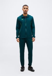 Άνδρας που στέκεται φορώντας σπορ σακάκι σκούρου τιρκουάζ και παντελόνι με μαύρα παπούτσια με δόντια, σε λευκό απλό φόντο.