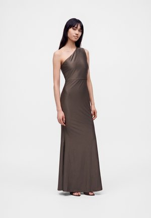 Femme portant une longue robe marron foncé asymétrique, debout, les bras détendus contre un fond clair uni.