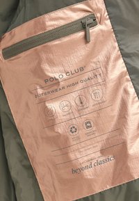 Metka odzieży wierzchniej Polo Club z brązową, metaliczną fakturą i nadrukowanymi ikonami pielęgnacji; zielone tło z tkaniny; detal kieszeni na suwak.