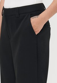 Black trousers with a smooth texture, featuring side pockets, a flat front, and a tailored fit. Subtle pleat detail near the waist.