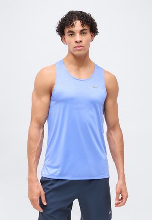 Uomo con capelli ricci che indossa una canotta sportiva azzurro chiaro e pantaloncini scuri, in piedi davanti a uno sfondo chiaro e uniforme.