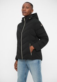 Doudoune noire avec un motif en chevrons, capuche et détails de fermeture éclair dorés. Comprend des poches latérales et une texture lisse et matelassée.