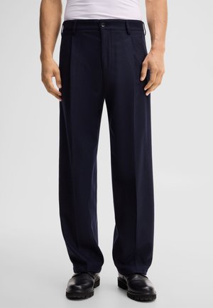 Uomo con pantaloni neri a gamba larga, scarpe nere e camicia bianca infilata, in piedi davanti a uno sfondo semplice.