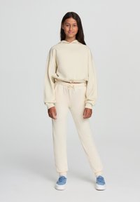 Felpa color crema con cappuccio e cordino, abbinata a pantaloni della tuta a gamba stretta coordinati. Texture del tessuto morbida, vestibilità comoda e polsini a costine.