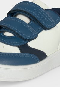 Gros plan sur une basket blanche et bleue avec deux brides Velcro bleues et des coutures visibles sur les brides et la chaussure.