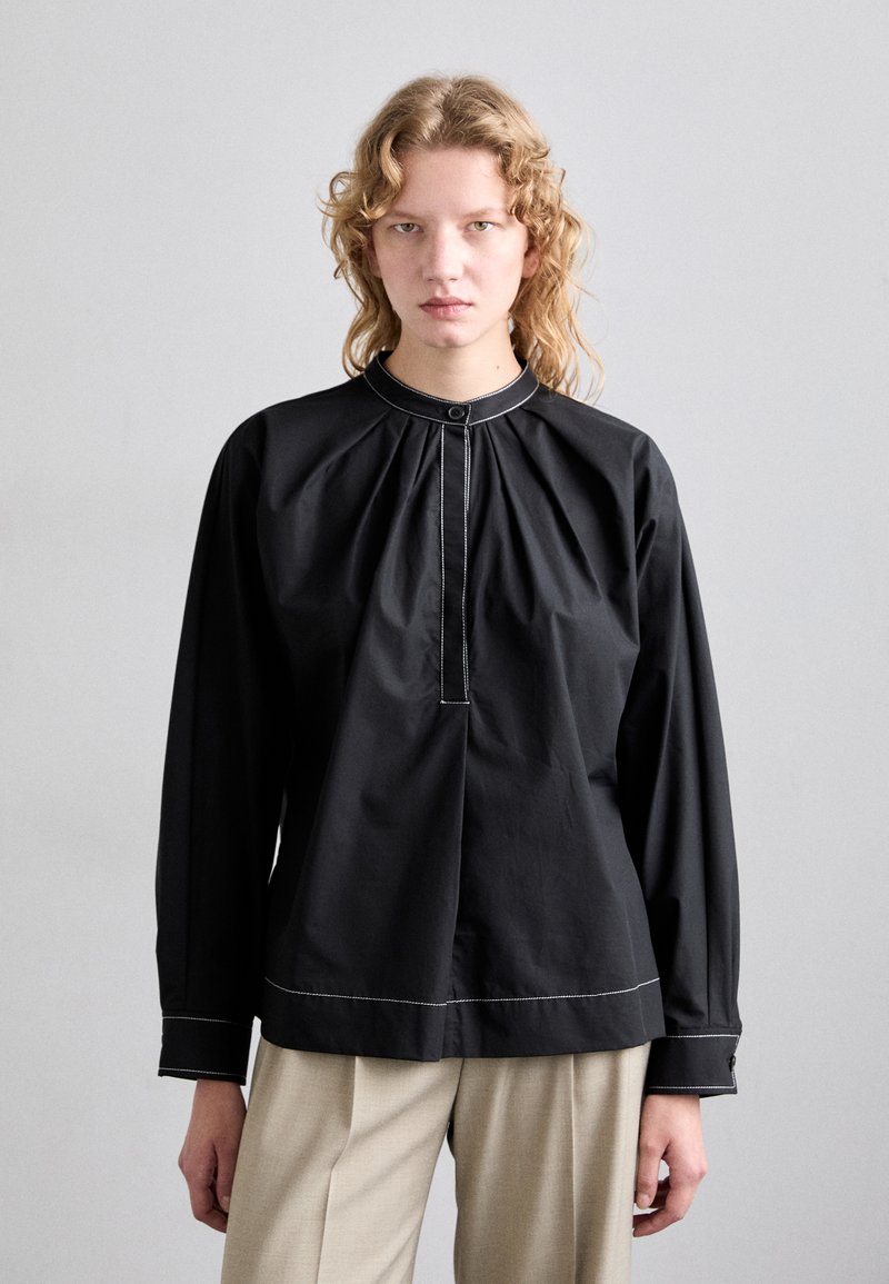 Blusa negra de manga larga con cuello fruncido, cierre de botones y costuras blancas contrastantes. Combinada con pantalones de color claro.