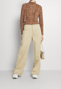 Braun langärmliges Oberteil mit kleinen weißen Blumenmustern, kombiniert mit weiten beigen Hosen. Weiße Sneakers und eine kleine beige Handtasche runden den Look ab.