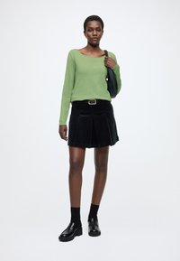 Kobieta w zielonym, długim rękawie, czarnej plisowanej spódnicy z paskiem, czarnych skarpetkach, mokasynach i z czarną torbą na ramię na jednolitym tle.