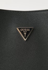Sac en cuir texturé noir avec un plaqué logo triangulaire en or et noir portant le nom de la marque "GUESS" et un design de point d'interrogation.
