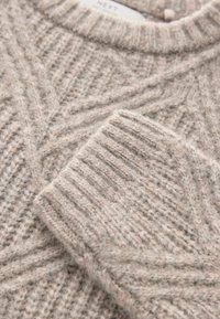 Beiger Strickpullover mit geripptem Kragen und Bündchen, versehen mit einem strukturierten V-Muster, das sich durch den gesamten Stoff zieht. Weiches, kuscheliges Material.