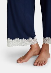 Blote voeten naast marineblauwe pyjamabroek met witte kanten bloemenrand aan de enkels op een witte achtergrond.