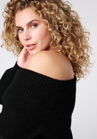 Zwarte off-the-shoulder trui met geribbelde textuur, voorzien van een brede halslijn. Model heeft krullend haar en poseert met haar hand bij haar nek.