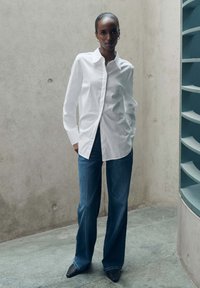 Witte overhemd met een klassieke kraag, gecombineerd met donkerblauwe wijde jeans en scherpe zwarte schoenen. Eenvoudig en strak ontwerp.