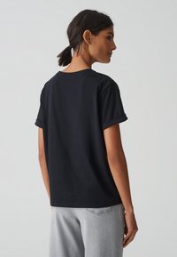 T-shirt nera in cotone con maniche arrotolate, vestibilità rilassata e scollatura rotonda. Modello che lo indossa con jeans grigi chiari a vita alta.
