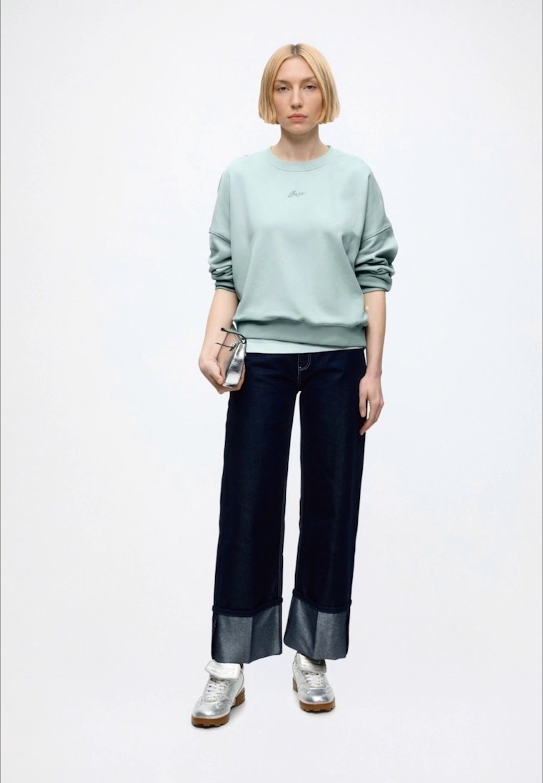 Femme aux cheveux blonds courts portant un sweat-shirt bleu clair, un jean large foncé avec des poignets métalliques, des baskets argentées et tenant un petit sac argenté.