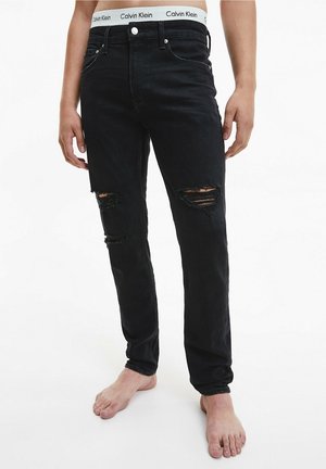 Man in zwarte gescheurde spijkerbroek en witte Calvin Klein-slip met zichtbare tailleband, blootsvoets staand tegen een witte achtergrond.