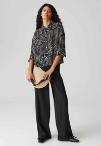 Camicia a righe in bianco e nero con una vestibilità ampia, abbinata a pantaloni neri a gamba larga. Tiene una borsa in paglia testurizzata con una tracolla nera.
