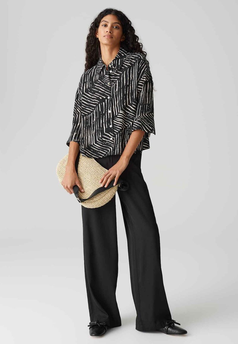 Czarna i biała koszula w wzory o luźnym kroju, w połączeniu z szerokimi czarnymi spodniami. Trzyma teksturowaną torbę ze słomy z czarnym paskiem.