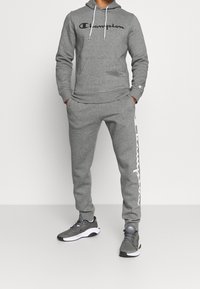 Felpa grigia Champion con logo nero, abbinata a pantaloni di tuta grigi con branding bianco lungo il lato, indossata con scarpe da ginnastica nere e grigie.