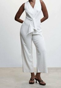 Mujer vestida con un mono sin mangas de rayas blancas y un detalle de nudo en el frente, con sandalias de tacón alto marrones, de pie con las manos detrás de la espalda.