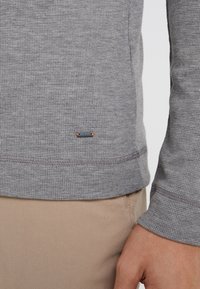 Graues, strukturiertes Langarmshirt mit einem dezenten Logo-Tag am unteren Saum, ausgestattet mit einem gerippten Kragen und Bündchen, kombiniert mit beigen Hosen.