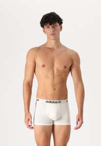 Weiße Boxershorts mit einem schwarzen Taillenbund, der das "adidas"-Logo und drei schwarze Streifen an den Seiten zeigt, aus einem leichten Stoff gefertigt.