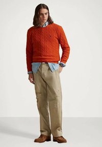 Młody mężczyzna z długimi włosami, ubrany w pomarańczowy sweter w warkocze, niebieską koszulę, beżowe spodenki z przetarciami i brązowe skórzane buty, stoi z ręką w kieszeni.