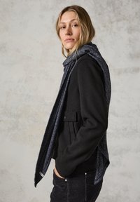 Sort uldjakke med en frontlomme, parret med et mørkeblåt struktureret tørklæde, begge med en blød finish. Model står sideværts med hænderne i lommerne.