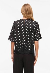 Blouse à pois noirs en tissu transparent, avec un bord en dentelle festonnée à l'ourlet et une fermeture à boutons dans le dos. Manches courtes et fluides.