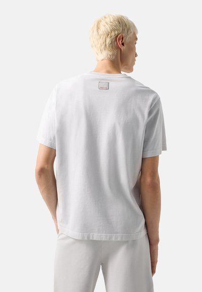 Personne aux cheveux blonds courts portant un t-shirt blanc uni à manches courtes et un pantalon blanc, vue de dos.