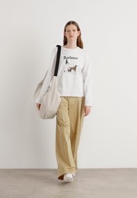 T-shirt blanc à manches longues avec un graphique "Barbour", pantalon beige à larges jambes, et un sac beige clair avec une bandoulière noire. Baskets blanches.