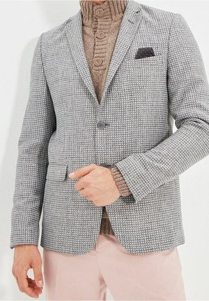 Mężczyzna w szarej, kraciastej marynarce na swetrze brązowym, zapinanym na guziki, w jasnoróżowych spodniach, poprawiający przód marynarki.